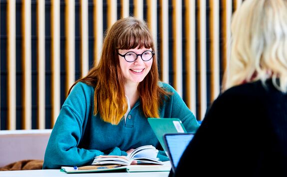 Two people sitting at a table facing each other. One has long hair and a teal shirt with an open book in front of them; the other has blonde hair and wears a black top. They appear to be in conversation or studying.